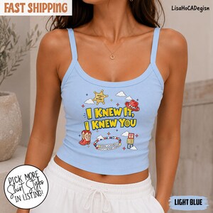 May include: A light blue ribbed tank top featuring a graphic print with the text "I KNEW IT, I KNEW YOU" in yellow, surrounded by illustrations of a sheriff badge, a cowboy hat, red cowboy boots, a beaded bracelet, and alphabet blocks.