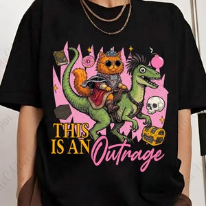 May include: Black t-shirt featuring a cartoon cat wearing a crown and sunglasses, riding a green dinosaur. The design includes a pink background with the text "THIS IS AN Outrage", a skull, a treasure chest, and other whimsical elements.