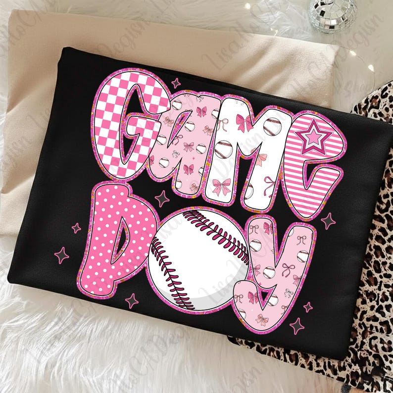 Może przedstawiać: Czarny t-shirt z napisem "Game Day" w kolorze r&oacute;żowym i białym. Litery mają r&oacute;żne wzory, w tym grochy, kratę i paski. Na środku znajduje się piłka do baseballu. Małe r&oacute;żowe gwiazdki otaczają tekst.