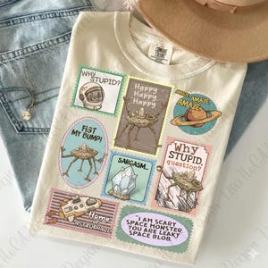 May include: Cream-colored t-shirt with various cartoon graphics and text. Designs include a space helmet, a planet with rings, and alien-like creatures. Text includes "Why Stupid?" and "Fist My Bump!"