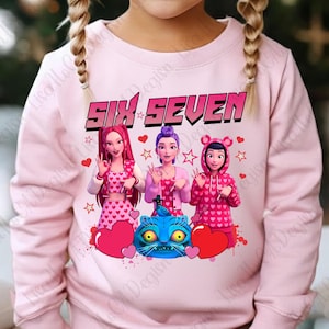 May include: A light pink sweatshirt featuring the text "SIX SEVEN" above a graphic of three cartoon girls. The girls are wearing pink outfits with heart patterns. A blue cat and red hearts are also featured in the design.