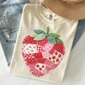 May include: Cream-colored t-shirt featuring a large, patchwork strawberry design. The strawberry is composed of various red and pink fabrics, including floral and gingham patterns. Green leaves and stem complete the design. The shirt is folded.