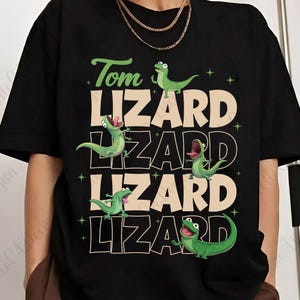 May include: Black t-shirt with a green and white graphic design. The design features the text "Tom Lizard" with several green cartoon lizards. The lizards are in various poses, with some having their mouths open.