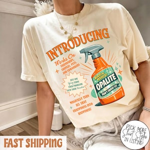 Könnte beinhalten: Cremefarbenes T-Shirt mit dem Wort "INTRODUCING" in Orange. Das Shirt zeigt eine Grafik einer orangefarbenen Sprühflasche mit der Aufschrift "OPALITE" und dem Text "Works On: Friendships, Couples, Pets, and Coworkers". Zusätzlicher Text lautet "Magical Results!" und "Don't Sweat It!"