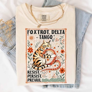 May include: Cream-colored t-shirt featuring a tiger-striped cat holding a red snake, surrounded by floral illustrations. The shirt has the text "Foxtrot, Delta, Tango" and "Resist, Persist, Prevail" in a decorative frame. A gold necklace rests on the shirt.