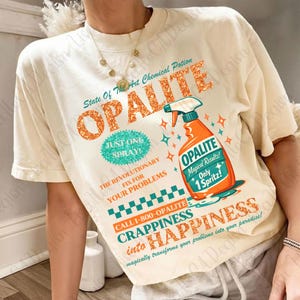 Könnte beinhalten: Cremefarbenes T-Shirt mit einem Grafikdesign im Retro-Stil. Das Design zeigt das Wort "OPALITE" in orangefarbenen Glitzerbuchstaben, eine Sprühflaschenillustration und Text, der für einen chemischen Trank wirbt. Das Shirt enthält den Text "Just One Spray!" und "Only 1 Spritz!"