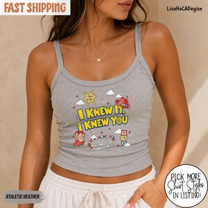 May include: A heather gray tank top featuring a graphic print with the text I KNEW IT, I KNEW YOU. The design includes a yellow sheriff star, a red cowboy hat, a red cowboy boot, a colorful beaded bracelet, and a stack of alphabet blocks.