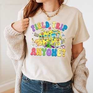 Può includere: Una maglietta beige con una grafica colorata di un autobus scolastico che vola nell'aria con il testo "Field Trip Anyone?" stampato su di essa.