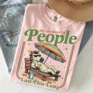 May include: Pink t-shirt with the text "My tolerance for People Last This Long". The shirt features a cartoon zebra relaxing on a beach chair under a rainbow umbrella, holding a drink. A bucket with a bottle of wine is next to the chair.