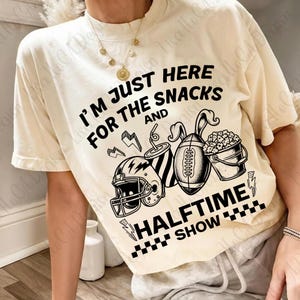 May include: Cream-coloured t-shirt with the text "I'M JUST HERE FOR THE SNACKS AND HALFTIME SHOW." The graphic includes a football helmet, a drink, a football, and a bucket of popcorn. Black and white design.