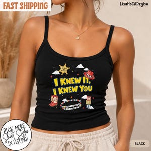 May include: A black ribbed tank top featuring a graphic print with the yellow text I KNEW IT, I KNEW YOU, a gold sheriff star, a red cowboy hat, a beaded bracelet, and stacked alphabet blocks, all surrounded by small white clouds and stars.