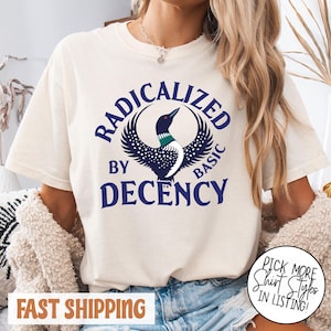 May include: Cream-colored t-shirt with a graphic design featuring a loon and the words "RADICALIZED BY DECENCY." The loon is depicted with a black head, a green neck, and a white and blue speckled body. The text is in navy blue.