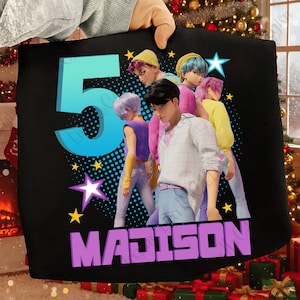 Peut inclure: Sac fourre-tout noir avec un groupe de personnages stylisés aux cheveux et vêtements colorés. Le chiffre "5" en bleu et le nom "MADISON" en violet sont également affichés. Des étoiles jaunes et une étoile blanche agrémentent le design.