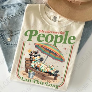 May include: Cream-colored t-shirt with the text "My tolerance for People Last This Long." The graphic features a zebra in sunglasses relaxing on a beach chair under a rainbow umbrella, holding a drink. The shirt has a Comfort Colors tag.