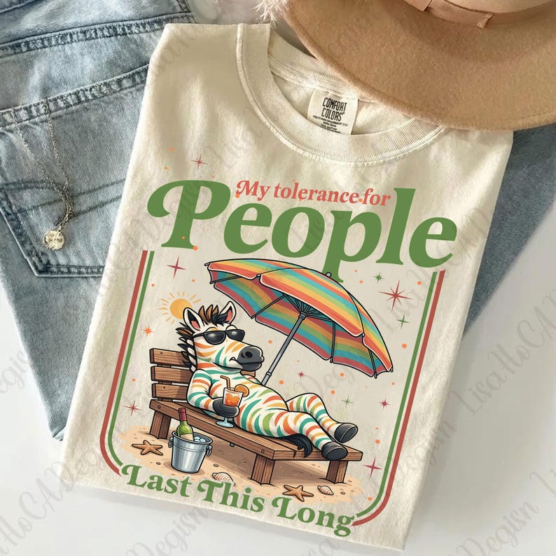 May include: Cream-colored t-shirt with the text "My tolerance for People Last This Long." The graphic features a zebra in sunglasses relaxing on a beach chair under a rainbow umbrella, holding a drink. The shirt has a Comfort Colors tag.