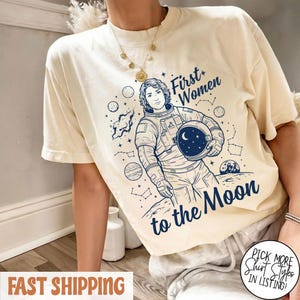 Könnte beinhalten: Cremefarbenes T-Shirt mit blauem Aufdruck einer Astronautin, die einen Helm mit Mond und Sternen hält. Der Text lautet "First Women to the Moon". Das Shirt hat kurze Ärmel und eine lockere Passform.