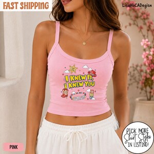 May include: A light pink ribbed tank top featuring a graphic print with the text I KNEW IT, I KNEW YOU. The design includes a yellow sheriff star, a red cowboy hat, a red cowboy boot, a colorful beaded bracelet, and a stack of alphabet blocks.