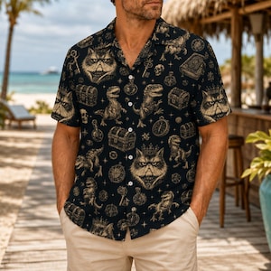 May include: A black short-sleeved button-down shirt featuring a repeating pattern of gold-toned illustrations including crowned cats, dinosaurs, treasure chests, keys, skulls, and compasses, with a notched collar and a front button closure.