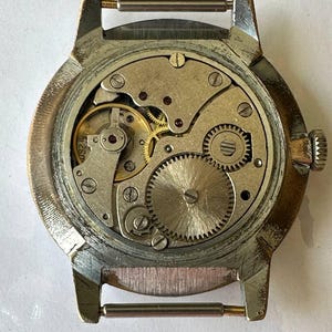May include: The back of a silver wristwatch with a round, silver case and a visible mechanical movement. The watch has a brown leather strap with a silver buckle.