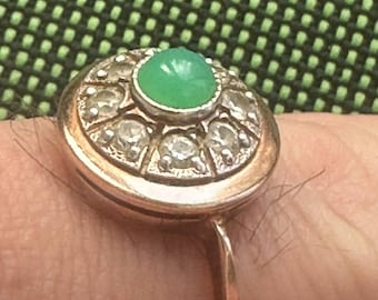 May include: A gold-toned ring featuring a central, round, green gemstone surrounded by a halo of smaller, clear gemstones. The ring's setting is detailed, and the band is slender and polished.