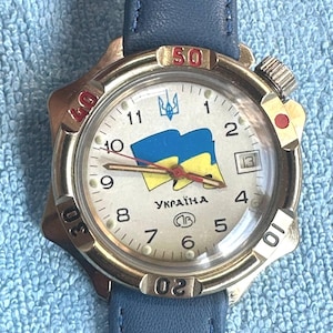Peut inclure: Une montre-bracelet avec un bracelet en cuir bleu et un boîtier en métal argenté. Le cadran blanc présente un drapeau bleu et jaune, le trident ukrainien, l'inscription "УКРАЇНА" et un guichet de date. La lunette tournante porte des chiffres rouges et noirs.