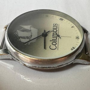 May include: A silver wristwatch with a white face and black hands. The face has a ship design and the word "Columbus" printed on it.
