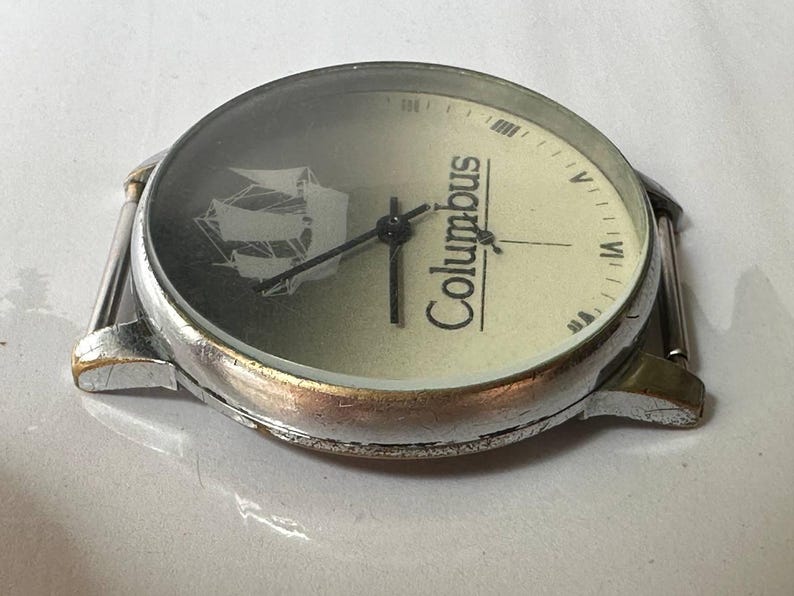 May include: A silver wristwatch with a white face and black hands. The face has a ship design and the word "Columbus" printed on it.