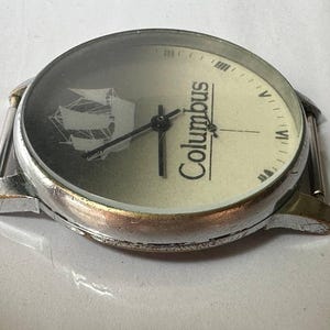 May include: A silver wristwatch with a white face and black hands. The face has a picture of a ship and the word "Columbus" printed on it.