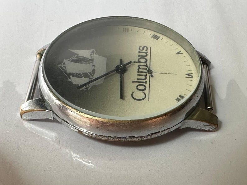 May include: A silver wristwatch with a white face and black hands. The face has a picture of a ship and the word "Columbus" printed on it.