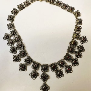 May include: A silver necklace with a delicate filigree design and red gemstones. The necklace has a pendant that hangs down in the center.