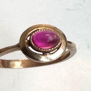 Può includere: Un anello color oro con una vibrante gemma magenta di forma ovale. La gemma è incastonata in una lunetta dettagliata e la fascia dell'anello ha un design semplice ed elegante. L'anello è mostrato su uno sfondo bianco.