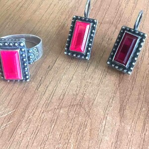 May include: A silver-toned ring and matching earrings set. The ring and earrings feature a rectangular, bright pink stone set within a beaded frame. The earrings are a drop style with a hook closure.