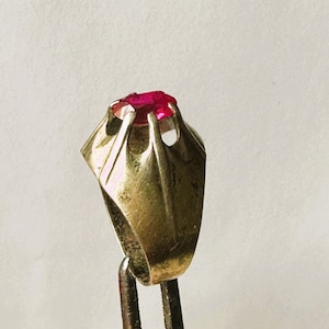 Può includere: Un anello d'oro con una pietra preziosa rossa incastonata in un design unico e intricato. L'anello è su un espositore.