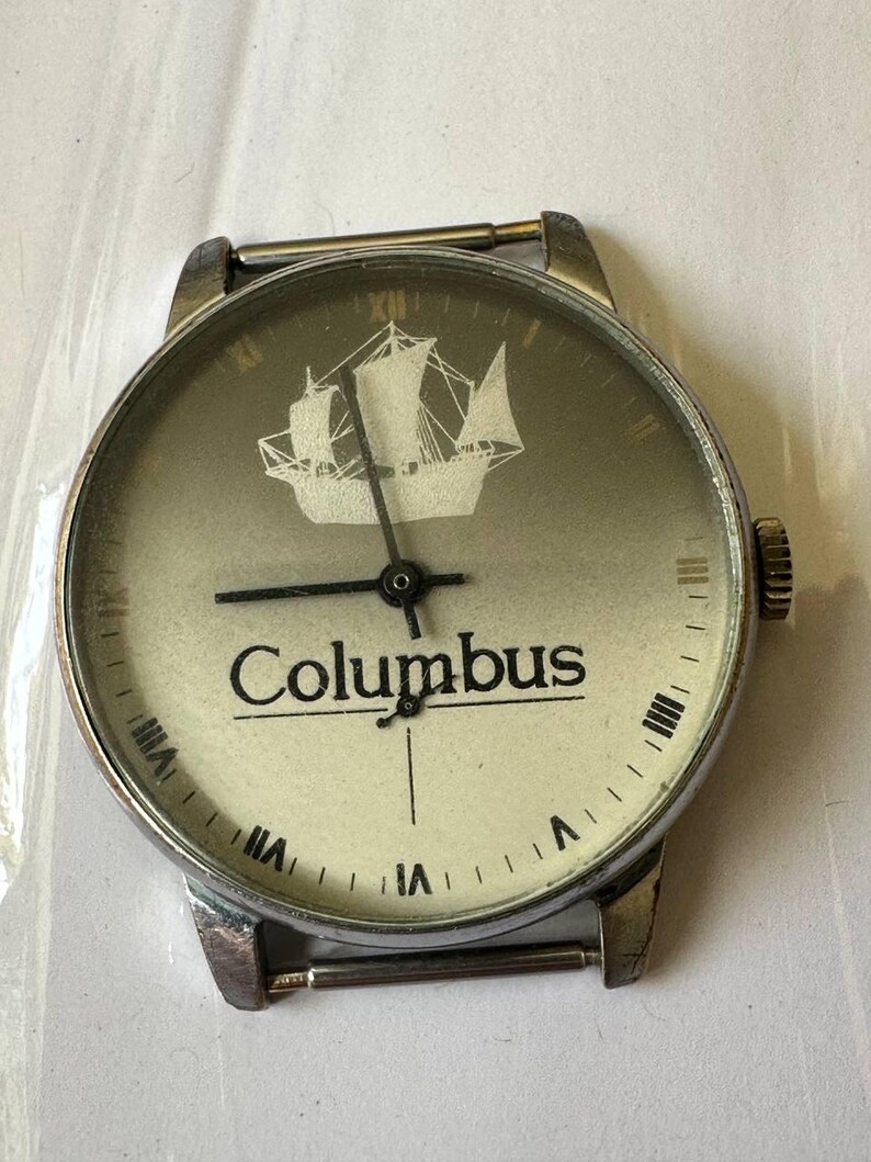 May include: A silver wristwatch with a white face and black hands. The face has a ship design and the word "Columbus" printed on it.