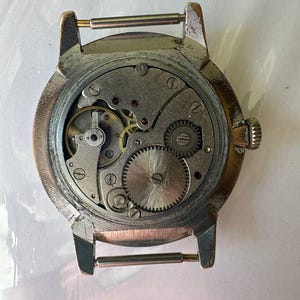 May include: The back of a silver wristwatch with a visible mechanical movement. The watch has a round case and a metal band.