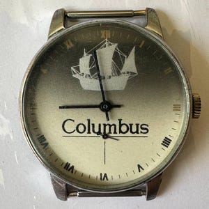 May include: A silver wristwatch with a white face and black hands. The face has a ship design and the word "Columbus" printed on it.