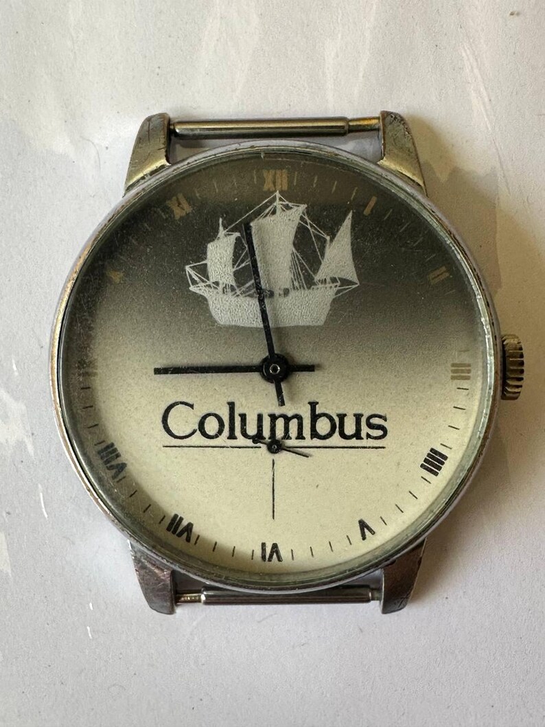 May include: A silver wristwatch with a white face and black hands. The face has a ship design and the word "Columbus" printed on it.