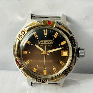Puede incluir: Un reloj de pulsera Vostok Century Time plateado y dorado con una esfera negra y agujas doradas. El reloj tiene un punto rojo en la parte superior y una correa de metal plateado.