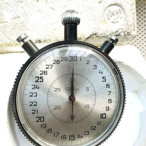 May include: A vintage stopwatch with a silver face and black bezel. The face displays numbers from 1 to 30, with a red second hand. The stopwatch has a black crown and two side buttons. The brand name is visible on the face.