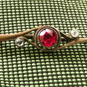 May include: A silver brooch with a red gemstone and two smaller clear gemstones. The brooch is in the shape of a curved line with a central oval.
