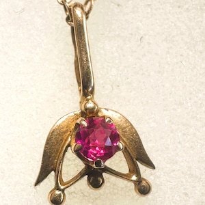 May include: A gold-toned pendant necklace featuring a vibrant, round, magenta gemstone. The pendant has a stylized floral design with curved leaves and small spherical accents. The chain is delicate and gold-colored.