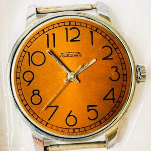 Peut inclure: Une montre-bracelet argentée avec un cadran rond orange. Le cadran de la montre comporte des chiffres noirs et un nom de marque 'Takema' noir. La montre a un bracelet en métal argenté.
