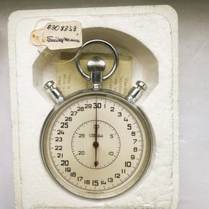 Op de afbeelding: Een vintage stopwatch met een zilveren kast en een witte wijzerplaat, met zwarte cijfers en markeringen. De stopwatch zit in een witte doos met een aangehecht label. Het label heeft het nummer "0308838" en Cyrillische tekst.