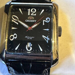 Può includere: Un orologio automatico Orient color oro con quadrante nero e numeri bianchi. L'orologio ha un cinturino in pelle nera ed è resistente all'acqua fino a 30 metri.