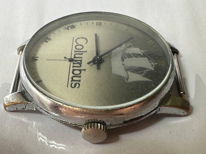 May include: A silver wristwatch with a white face and black hands. The face has the word "Columbus" printed on it and a ship design. The watch has a silver band.