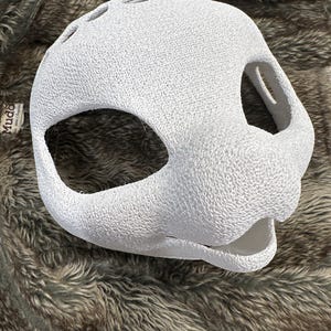 May include: A white, textured mask with large eye openings and a snout. The mask has several small holes on top and is made of a material that appears to be woven or knitted. The mask is designed to fit over the head.