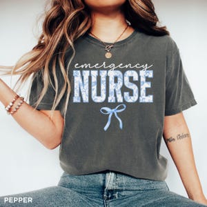 Puede incluir: Una camiseta gris oscuro con el texto "emergency nurse" en blanco con un patr&oacute;n floral azul. Un lazo azul est&aacute; debajo del texto.