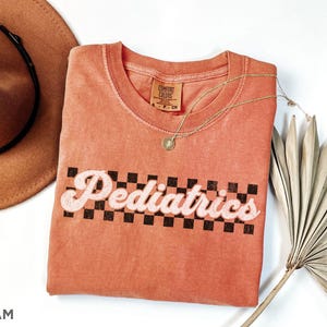 May include: A coral coloured t-shirt with a black and white chequered design and the text "Pediatrics" in a retro font.