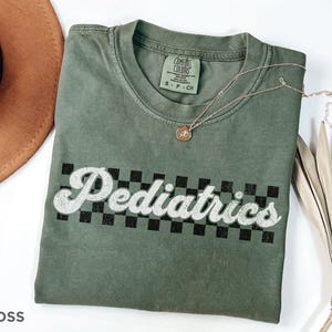 May include: A green t-shirt with a white graphic that says "Pediatrics" in a retro font with a black and white checkered pattern behind the text.