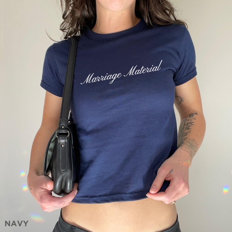 May include: Navy blue t-shirt with the text "Marriage Material" in a cursive font. The shirt has short sleeves and a crew neck. The word "NAVY" is at the bottom of the image.